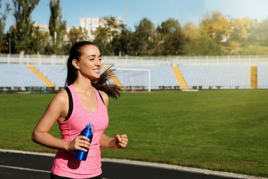 Photo of Sporty woman running with bottle of water at stadium on sunny day. Space for text