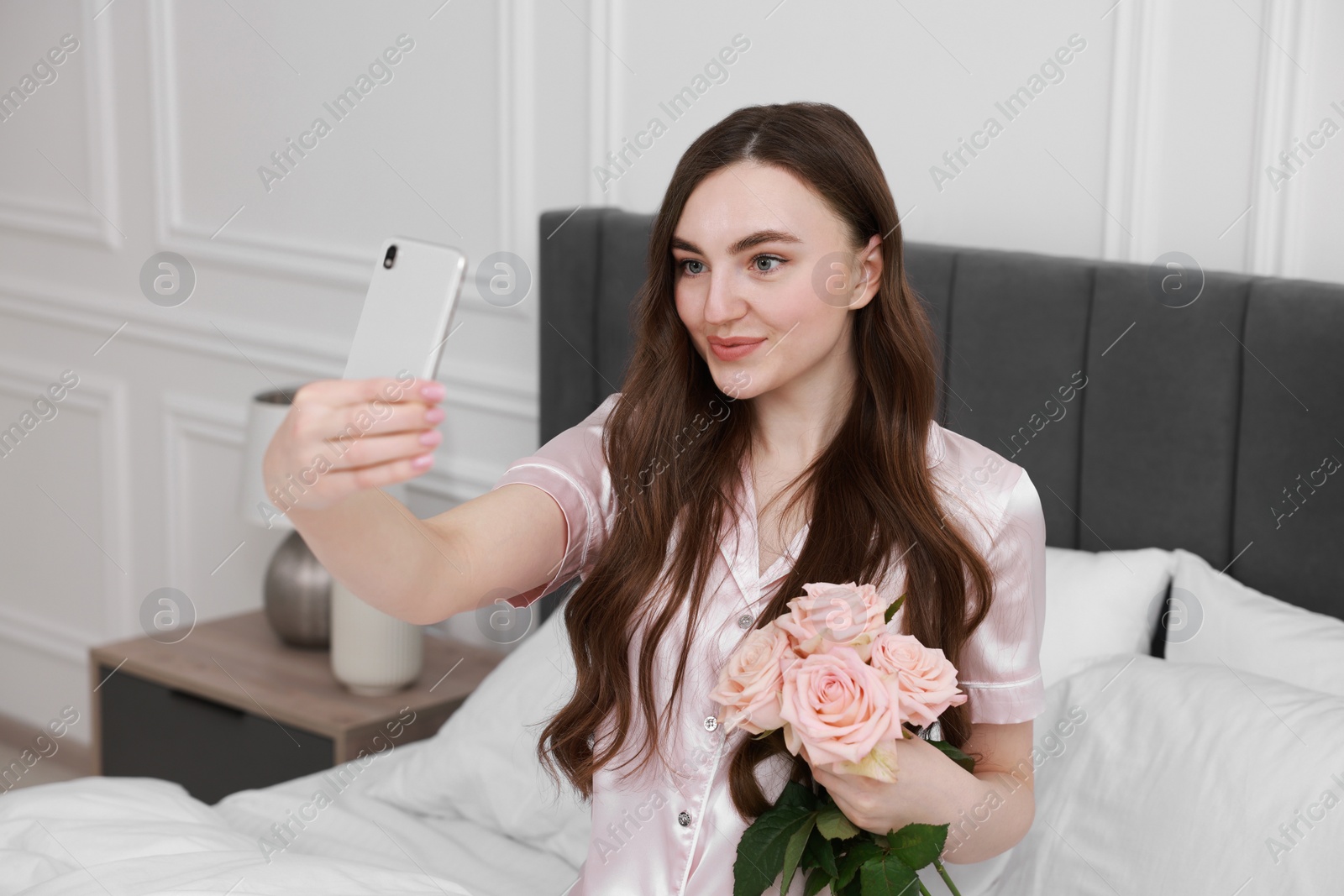 Photo of Beautiful young woman taking selfie with rose flowers on bed in room