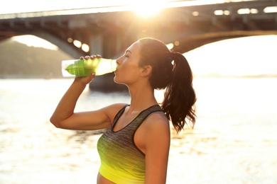Photo of Young woman drinking water after running near river on sunny day. Sports hydration