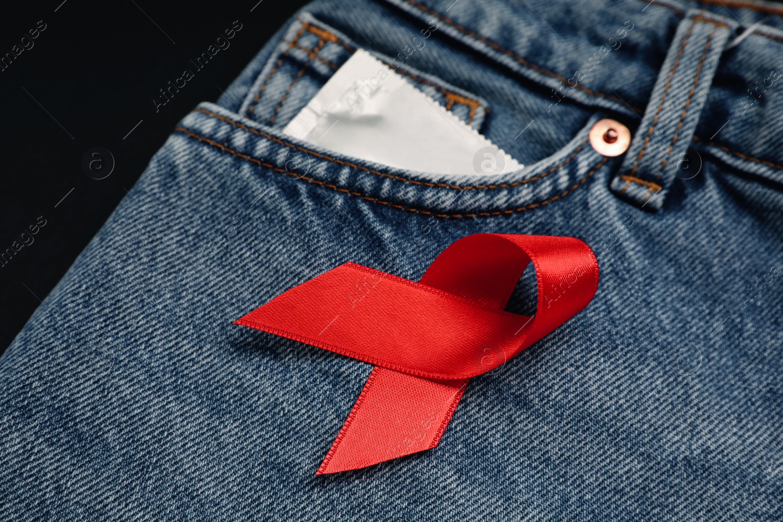 Photo of Condom and red ribbon in pocket of jeans on black background, closeup. AIDS disease awareness