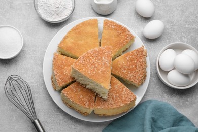 Pieces of tasty sponge cake, whisk and ingredients on light grey table, flat lay