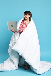 Happy woman in pyjama wrapped in blanket holding laptop on light blue background