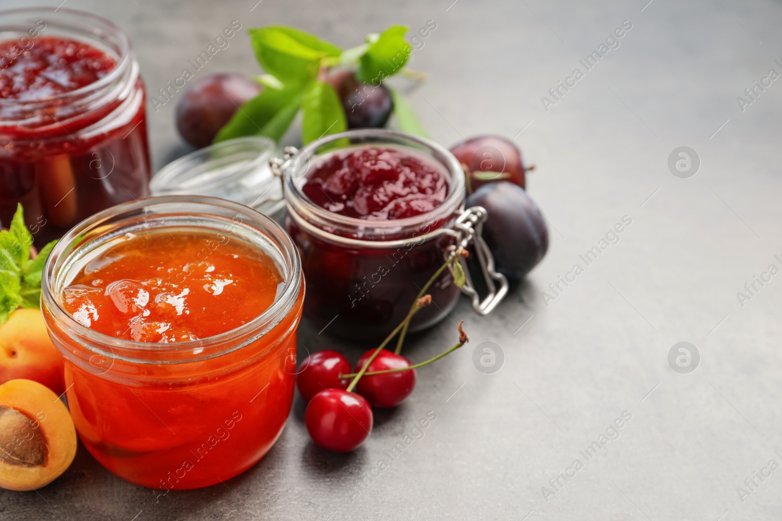 Photo of Jars with different jams and fresh fruits on grey table. Space for text