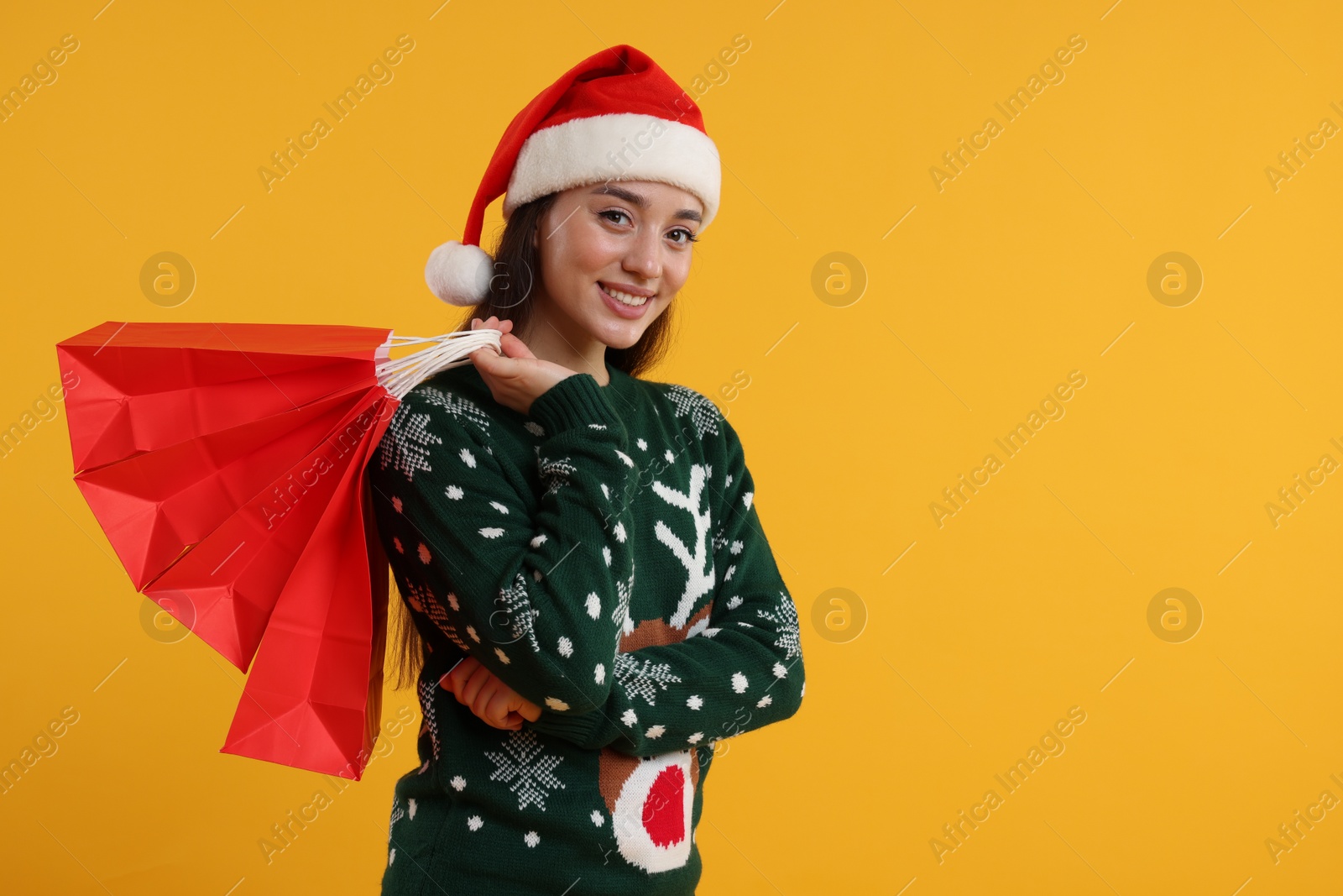 Photo of Happy young woman in Christmas sweater and Santa hat with shopping bags on orange background. Space for text