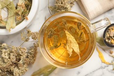 Photo of Freshly brewed tea and dried herbs on white marble table, flat lay