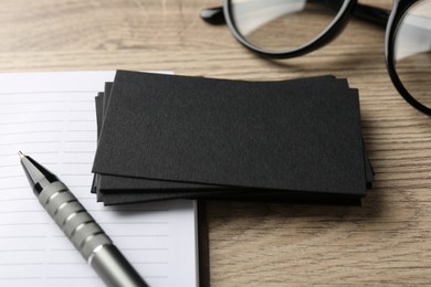 Photo of Blank black business cards, notebook, glasses and pen on wooden table, closeup. Mockup for design