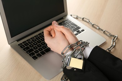 Woman showing chained hands near laptop at wooden table, closeup. Internet addiction