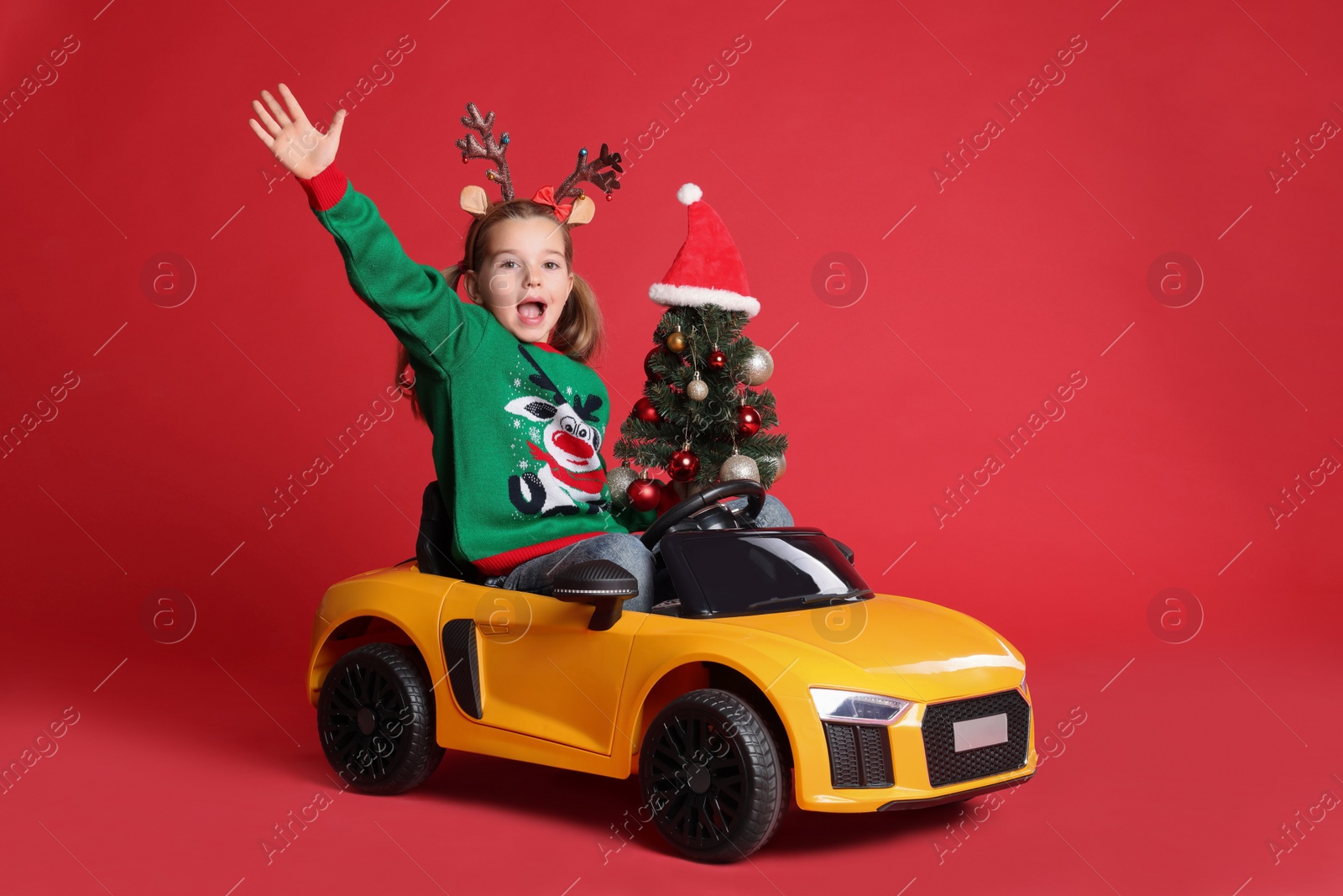 Photo of Cute little girl with Christmas tree driving children's electric toy car on red background