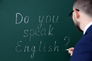 Photo of Teacher writing words Do You Speak English? on green chalkboard, closeup