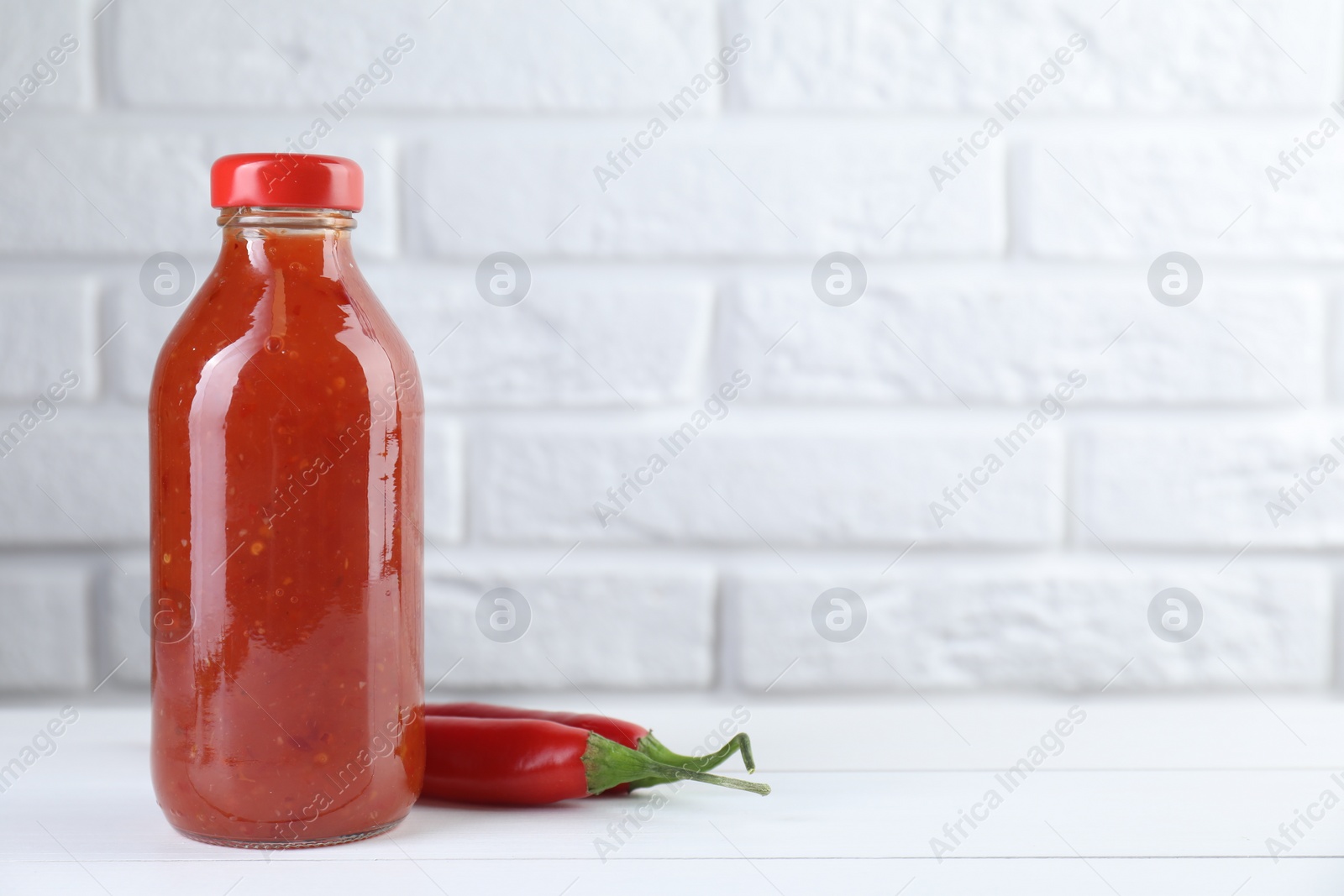 Photo of Spicy chili sauce in bottle and peppers on white wooden table, space for text