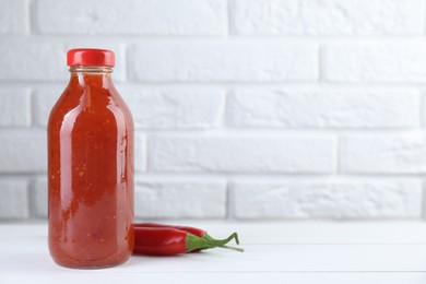 Photo of Spicy chili sauce in bottle and peppers on white wooden table, space for text