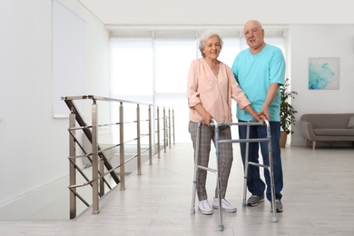 Photo of Elderly man helping his wife with walking frame indoors