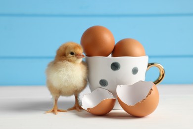 Photo of Cute chick with cup, eggs and pieces of shell on white wooden table, closeup. Baby animal