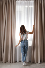 Woman opening window curtains at home in morning, back view