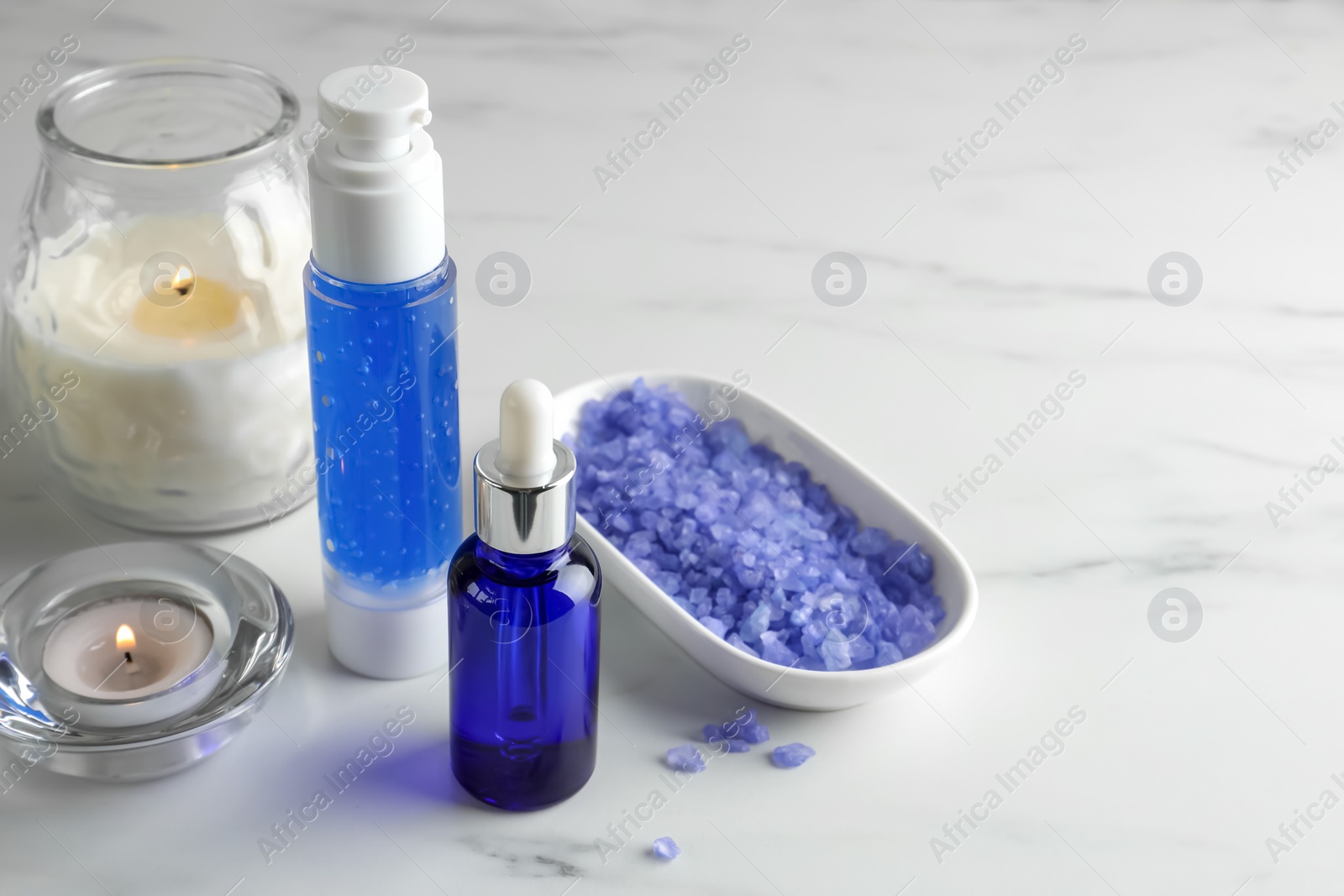 Photo of Spa composition. Bottles of cosmetic products, sea salt and burning candles on white marble table, space for text