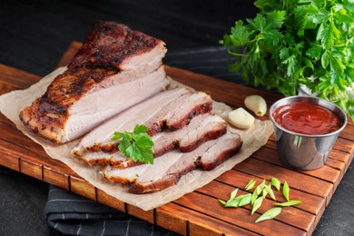 Photo of Pieces of baked pork belly served with sauce and parsley on black textured table, closeup