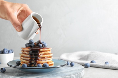 Woman pouring chocolate syrup onto fresh pancakes with blueberries at grey table, closeup. Space for text