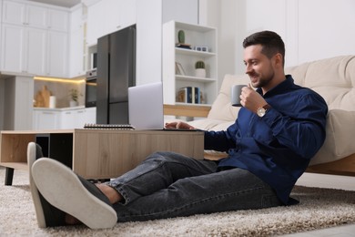 Happy man with cup of drink working on laptop at wooden coffee table in room