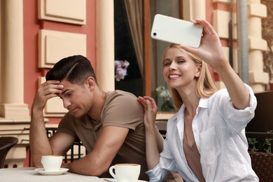 Young woman trying to take selfie with her displeased boyfriend in outdoor cafe. Boring date