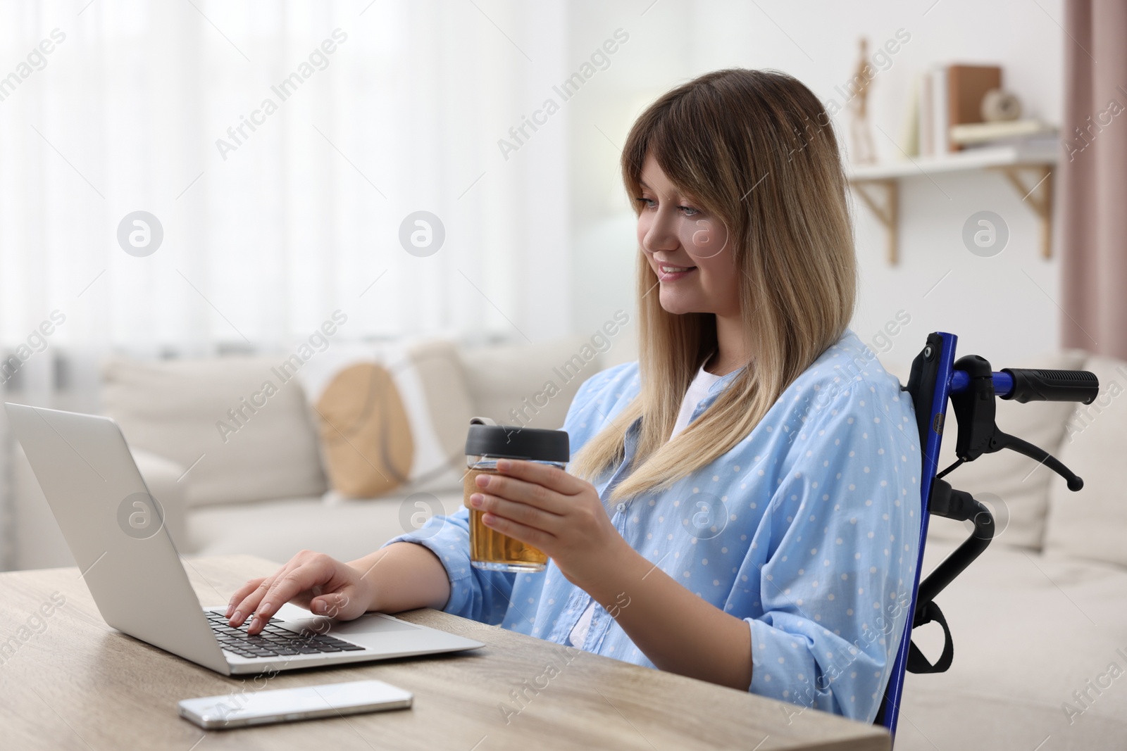 Photo of Woman in wheelchair with cup of drink using laptop at table in home office