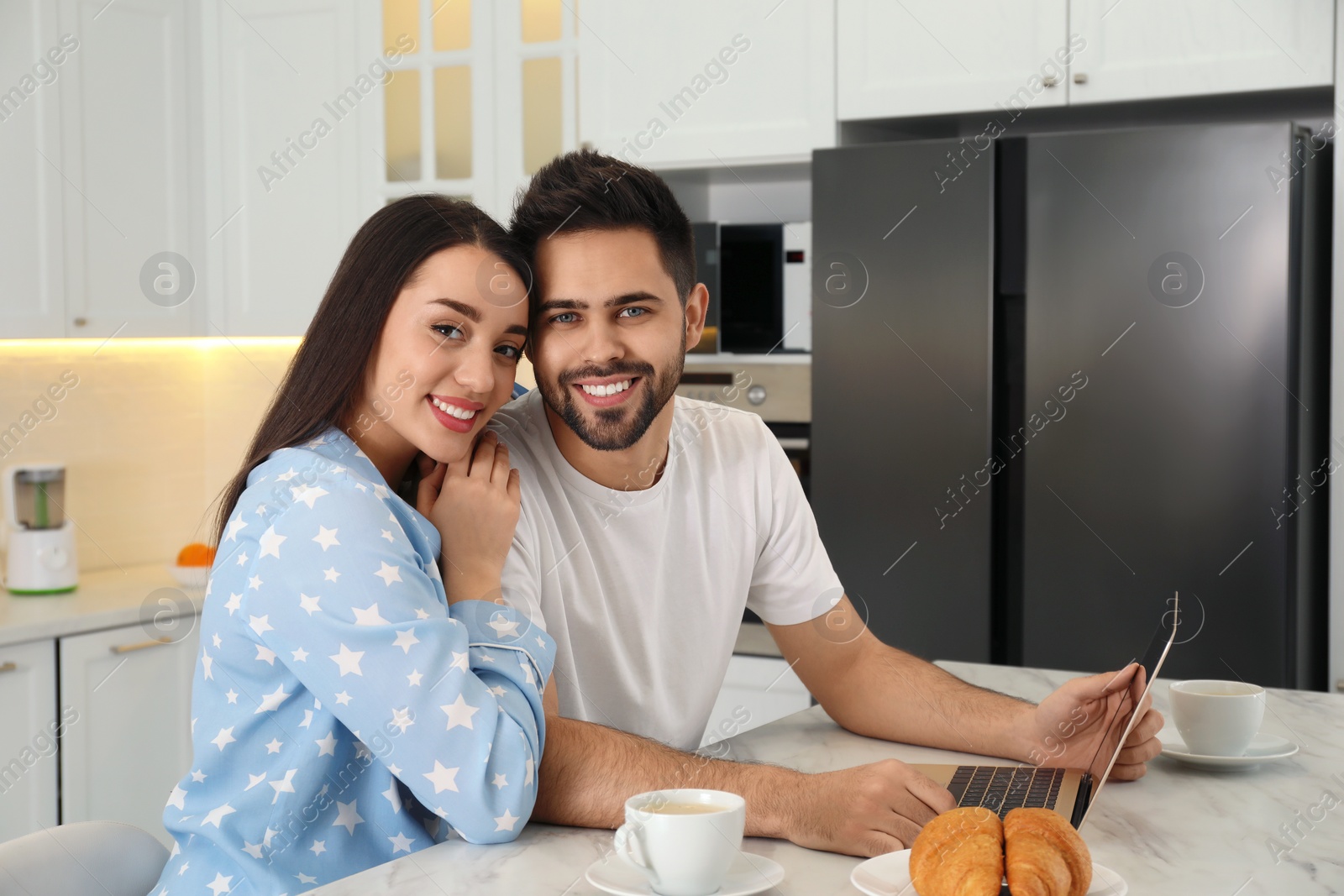 Photo of Happy couple in pajamas with laptop at kitchen table