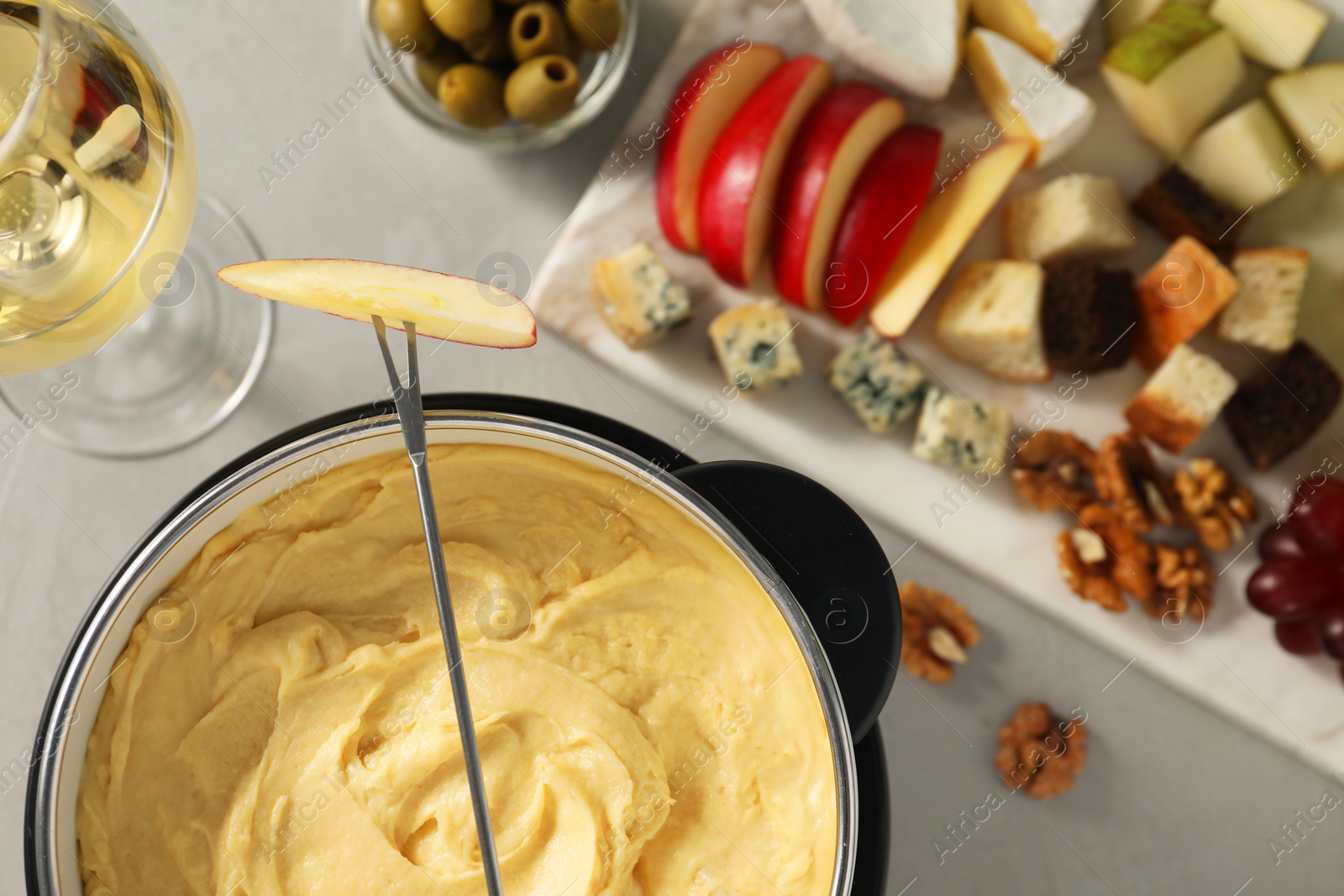 Photo of Fork with piece of apple, melted cheese in fondue pot, wine and snacks on grey table, flat lay