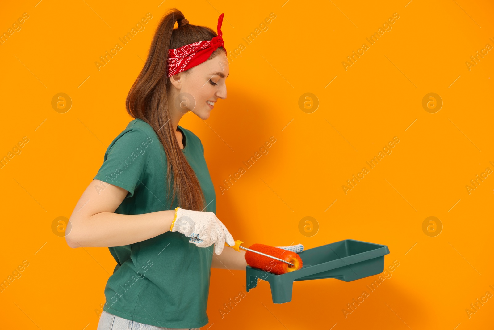 Photo of Happy designer with roller taking paint from container near orange wall