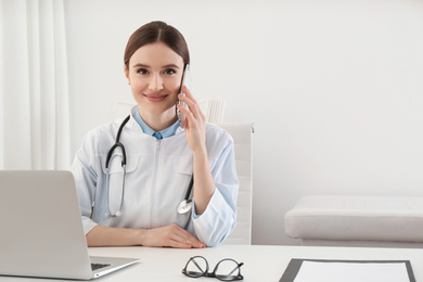 Young female doctor talking on phone at table in office
