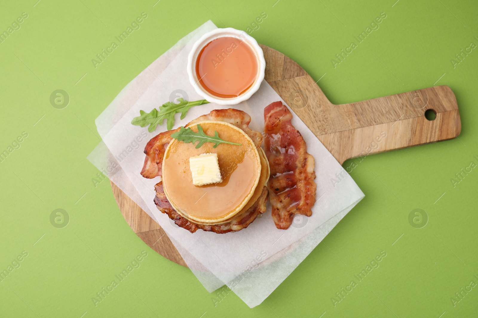 Photo of Delicious pancakes with bacon, butter, arugula and honey on green table, top view