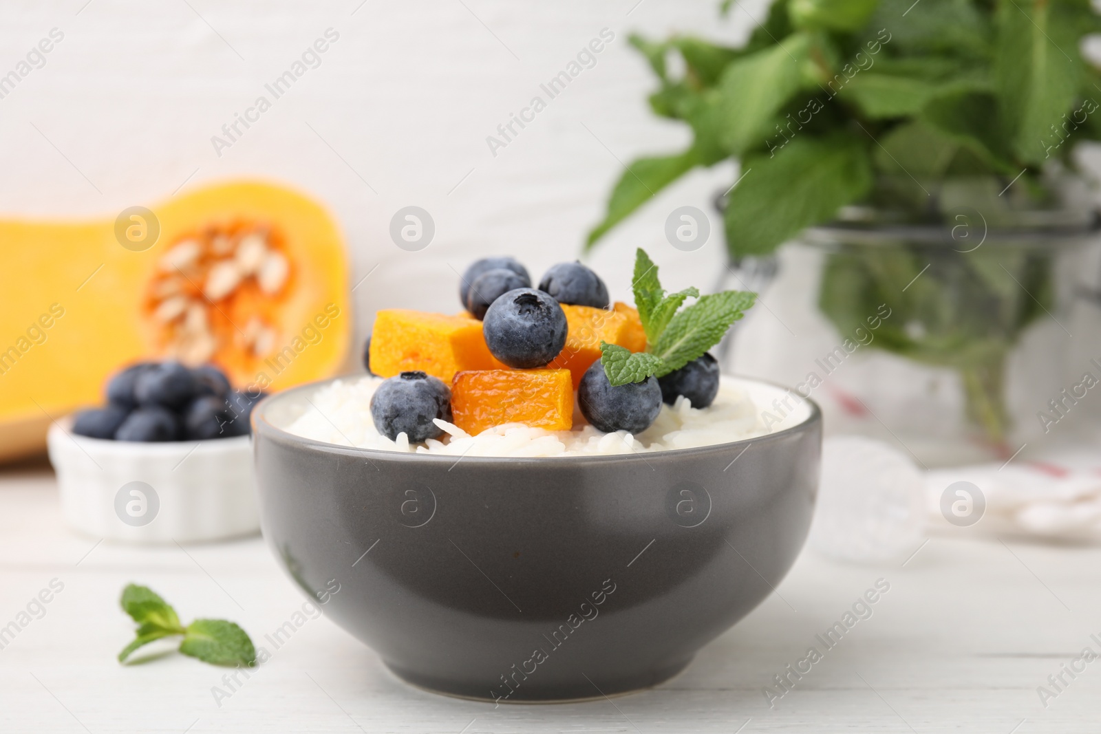 Photo of Bowl of delicious rice porridge with blueberries, pumpkin and mint on white table, closeup