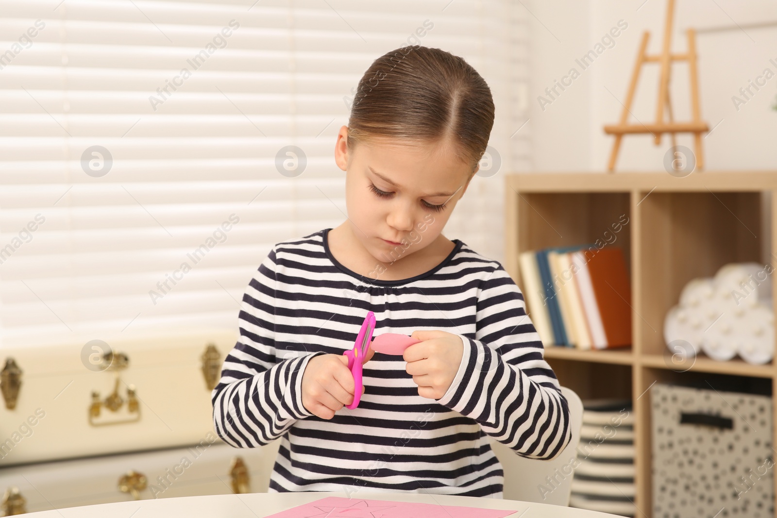 Photo of Cute little girl cutting pink paper at desk in room. Home workplace