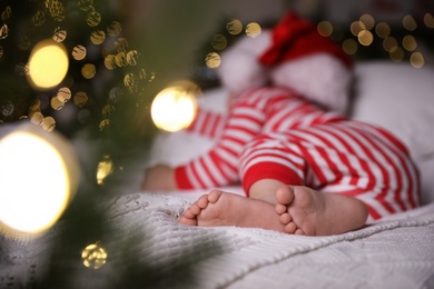 Photo of Baby in Christmas pajamas and Santa hat sleeping on bed indoors, focus on feet