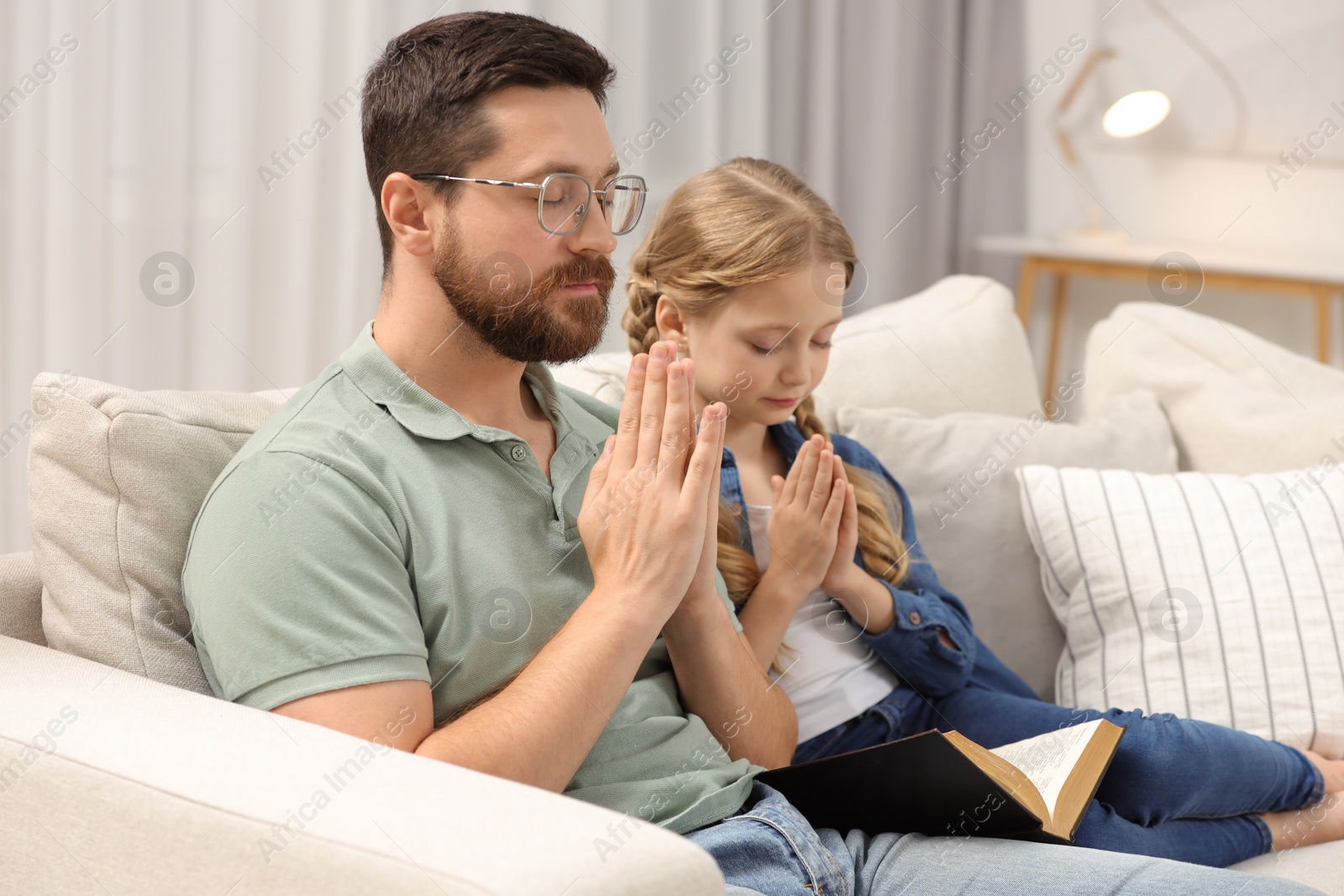 Photo of Girl and her godparent praying over Bible together on sofa at home