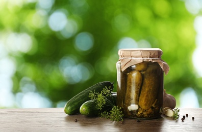 Image of Jar of tasty pickled cucumbers on wooden table against blurred green background. Space for text