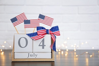 Photo of Wooden calendar with USA flags and bow on table against brick wall, space for text. Happy Independence Day