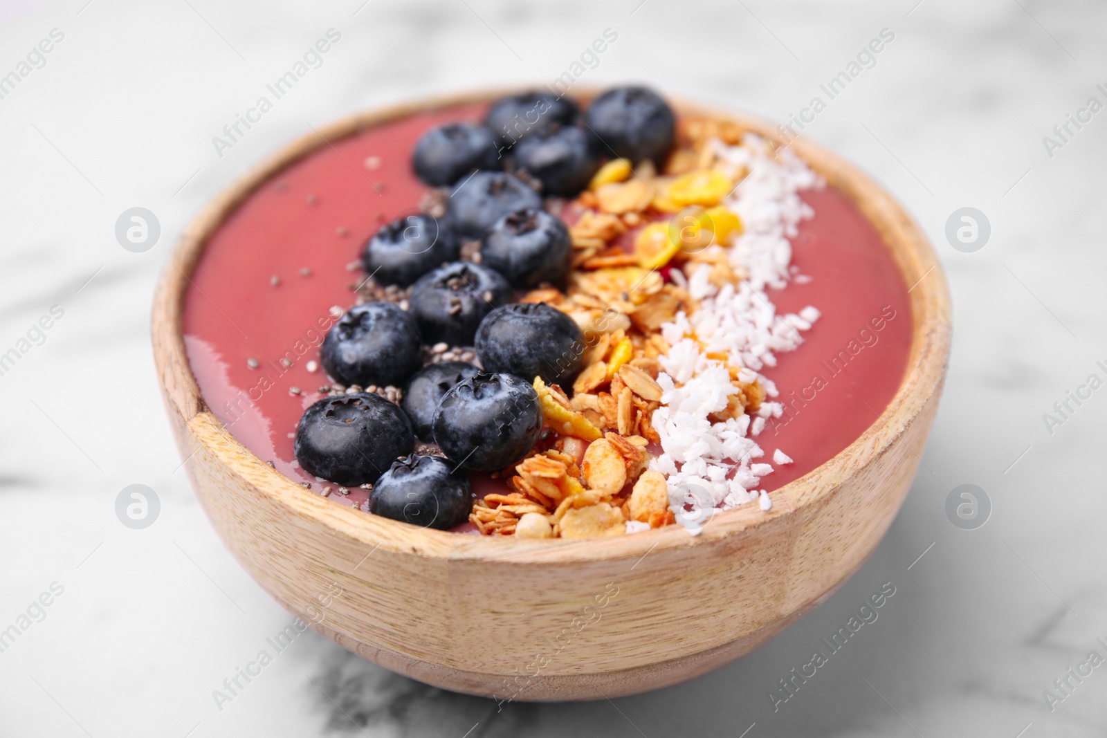 Photo of Bowl of delicious fruit smoothie with fresh blueberries, granola and coconut flakes on white marble table, closeup