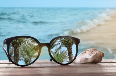 Image of Palms mirroring in sunglasses on wooden desk with seashell at sandy beach