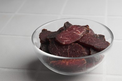 Photo of Bowl with pieces of delicious beef jerky on white tiled table, closeup