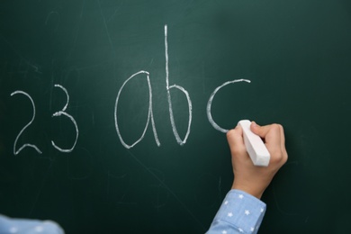 Photo of Little child writing letters and numbers on chalkboard, closeup