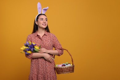 Photo of Happy woman in bunny ears headband holding wicker basket with painted Easter eggs and bouquet of flowers on orange background. Space for text