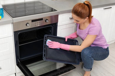 Photo of Woman cleaning oven tray with rag in kitchen