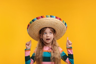 Photo of Cute girl in Mexican sombrero hat pointing at something on orange background