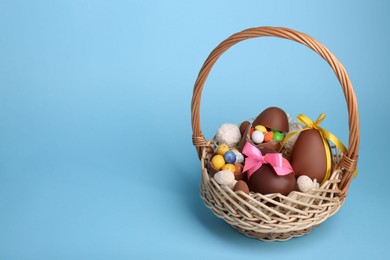 Photo of Wicker basket with tasty chocolate Easter eggs and different candies on light blue background, space for text