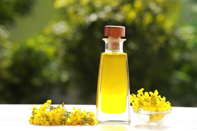 Photo of Rapeseed oil in glass bottle and yellow flowers on white wooden table outdoors