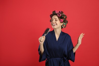 Photo of Beautiful young woman in silk bathrobe with hair curlers singing into hairbrush on red background