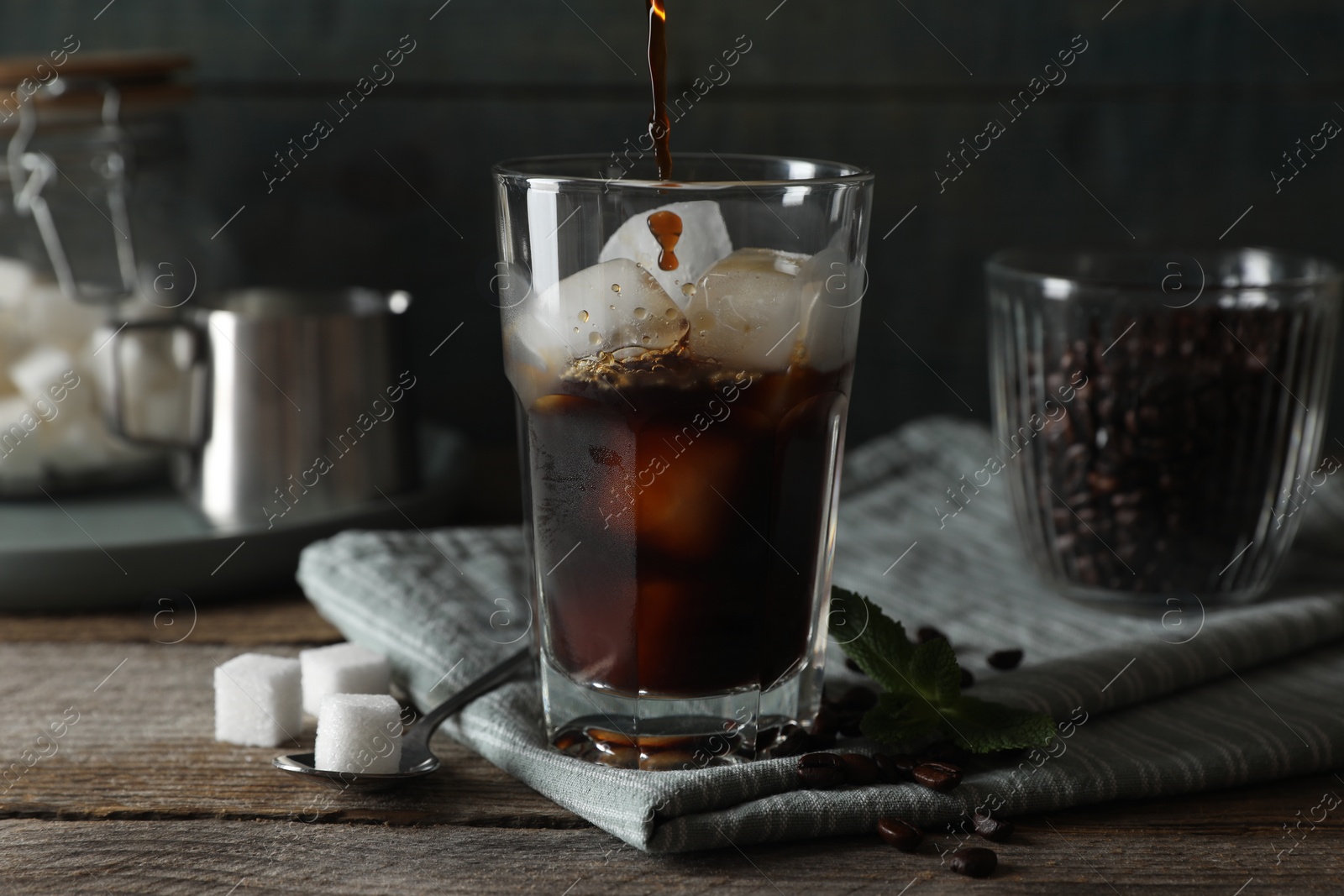 Pouring coffee into glass with ice cubes, mint and sugar on wooden table Photo of Pouring coffee into glass with ice cubes, mint and sugar on wooden table