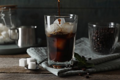 Photo of Pouring coffee into glass with ice cubes, mint and sugar on wooden table