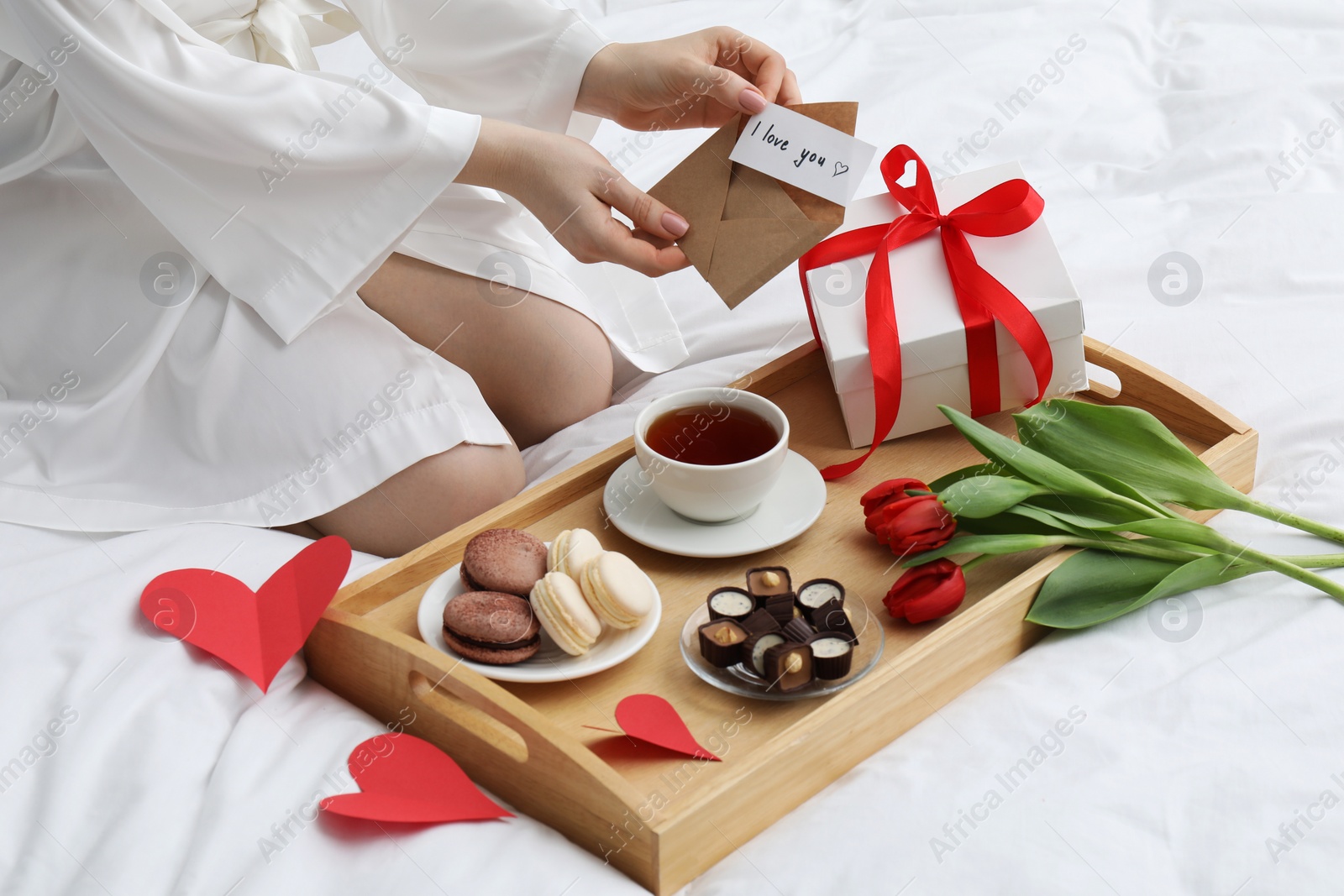 Photo of Tasty breakfast served in bed. Woman with I Love You card, desserts, tea, flowers and gift box at home, closeup