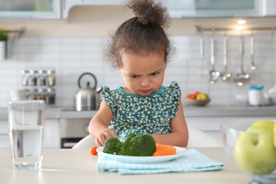 Photo of Cute African-American girl with plate of vegetables at table in kitchen