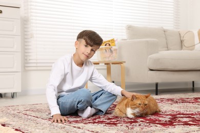Photo of Little boy petting cute ginger cat on carpet at home